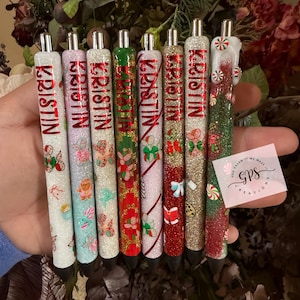 May include: A collection of personalized pens with the name "Kristin" in red lettering. The pens feature various designs, including glitter, gingerbread men, bows, and candy canes. Each pen has a silver tip and a black ink cartridge.