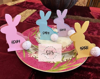 May include: Four pastel colored bunny rabbit place card holders with names written on them. The bunnies are sitting on white pom poms. The names are Henry, Emily, Hanna, and Gagan.