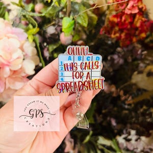 May include: A rectangular badge reel with the text "OHHH... THIS CALLS FOR A SPREADSHEET" in a colorful design. The badge reel has a clear plastic retractable cord and a metal clip. The background includes flowers and greenery.