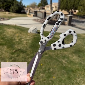 May include: Scissors with a white handle featuring black spots and a colorful design. The blades are silver. The scissors are held in front of a grassy background with a blurred view of trees and a house.