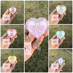 May include: A collection of heart-shaped badge reels with various designs. Each reel features a unique illustration, including boots, a skull, a crown, and books. The reels are held in a hand against a green background.