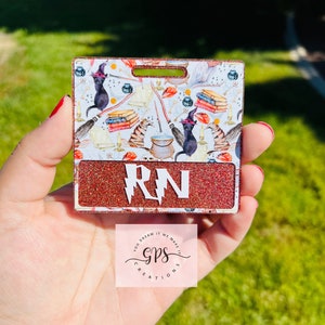 May include: A red glitter badge with a white background and a black cat, books, and a cauldron design. The badge has a white lightning bolt and the letters "RN" in black. The badge is held in a hand. The text "You Dream It We Make It GPS Creations" is visible below the badge.