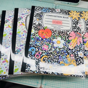 May include: A stack of composition notebooks with black spines and clear plastic covers. The notebooks feature a black and white speckled design with colorful illustrations of an apple, flowers, pencils, and the text "COMPOSITION BOOK".