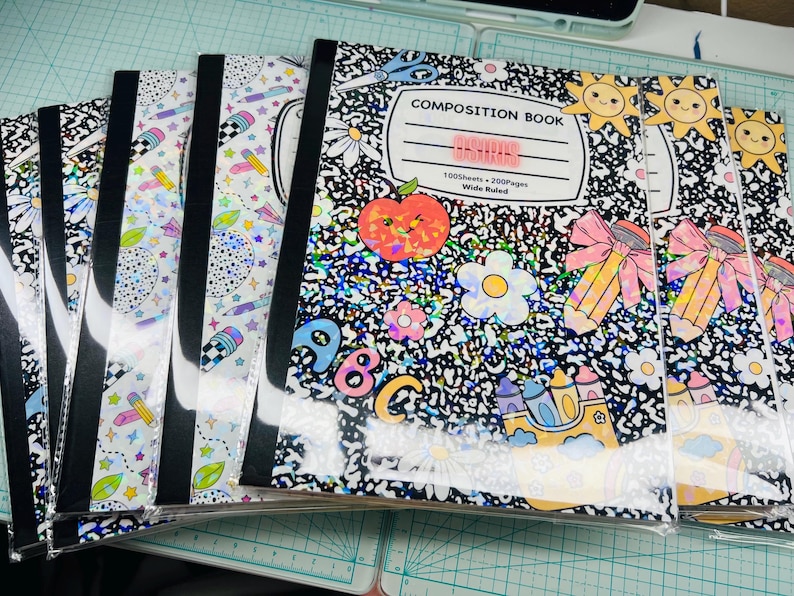 May include: A stack of composition notebooks with black spines and clear plastic covers. The notebooks feature a black and white speckled design with colorful illustrations of an apple, flowers, pencils, and the text "COMPOSITION BOOK".