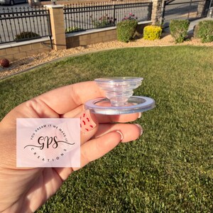 May include: A clear, plastic bottle stopper with a spiral design. The stopper is held in a hand against a background of green grass and a residential neighborhood. The text "YOU DREAM IT WE MAKE IT GPS CREATIONS" is visible.