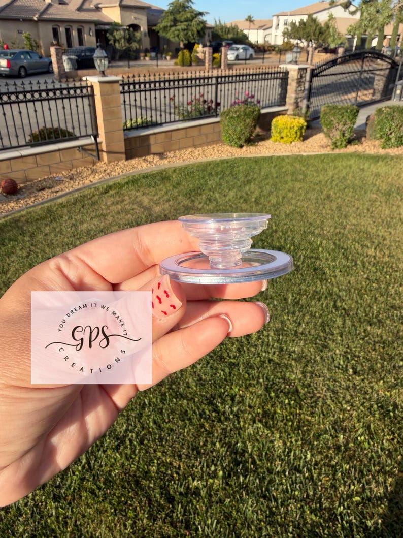 May include: A clear, plastic bottle stopper with a spiral design. The stopper is held in a hand against a background of green grass and a residential neighborhood. The text "YOU DREAM IT WE MAKE IT GPS CREATIONS" is visible.