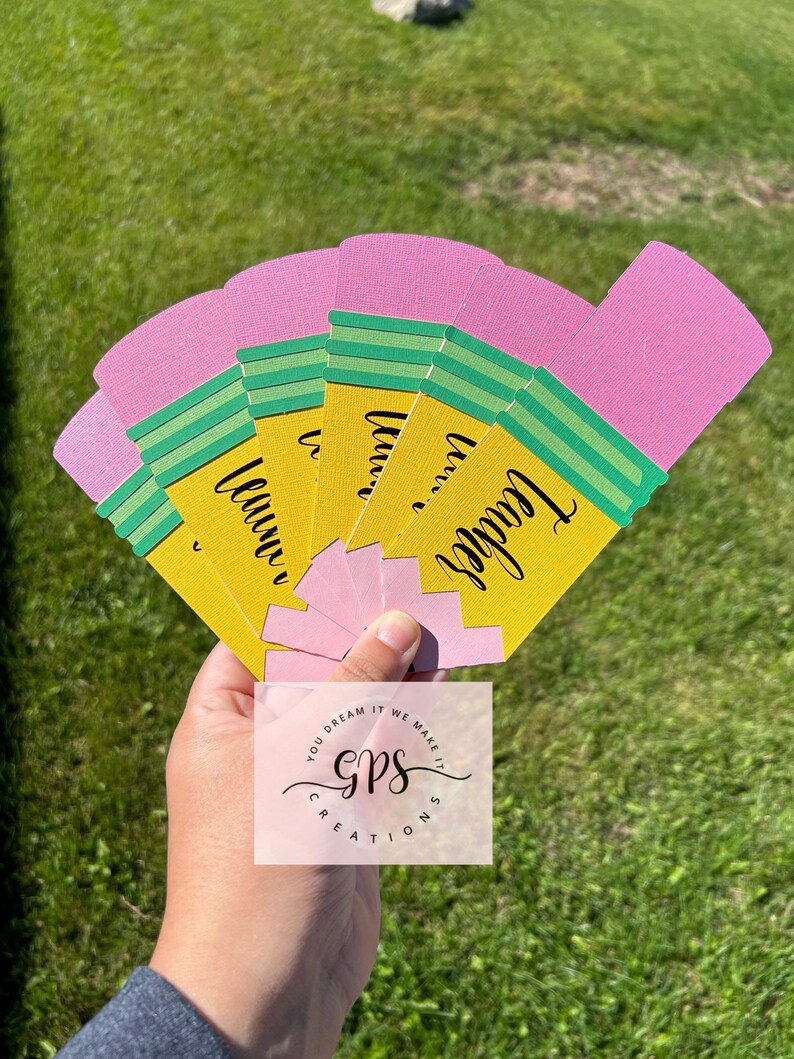 Pu&ograve; includere: Un set di sei segnalibri a forma di matita rosa e gialla con strisce verdi e la parola "Teacher" scritta su ogni segnalibro. I segnalibri sono tenuti in una mano. L'immagine include anche un logo per "GPS Creations" nella parte inferiore centrale dell'immagine.