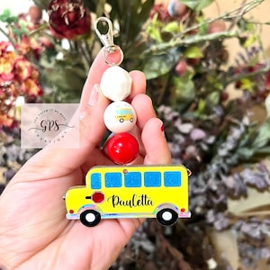 May include: A yellow school bus keychain with the name "Pauletta" on the side. The keychain has a silver clip and is decorated with white, yellow, and red beads.
