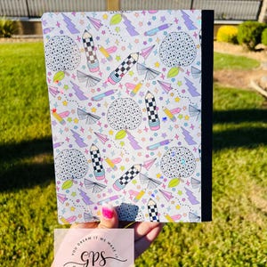 May include: A composition notebook with a white cover featuring a colorful pattern of pencils, stars, paper airplanes, and dotted circles. The notebook has a black spine and is held in front of a green background. The text "YOU DREAM IT WE MAKE IT GPS CREATIONS" is visible.