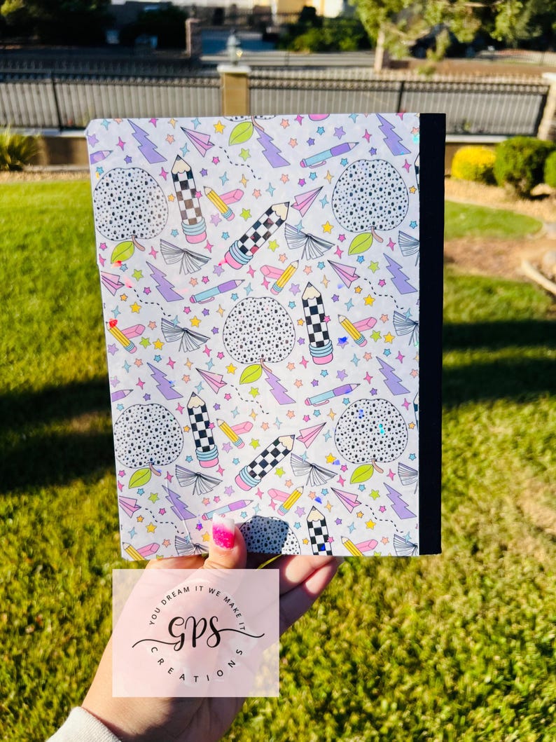 May include: A composition notebook with a white cover featuring a colorful pattern of pencils, stars, paper airplanes, and dotted circles. The notebook has a black spine and is held in front of a green background. The text "YOU DREAM IT WE MAKE IT GPS CREATIONS" is visible.