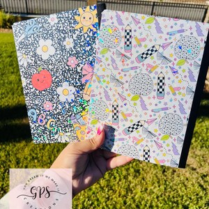 May include: Two notebooks with colorful, patterned covers are held up. One has a black background with white flowers, a smiling sun, and an apple. The other has a white background with pencils, stars, and lightning bolts. The notebooks are held in front of a green lawn.