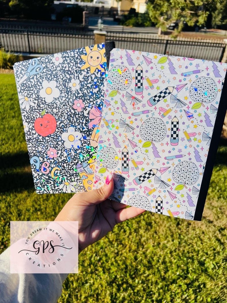 May include: Two notebooks with colorful, patterned covers are held up. One has a black background with white flowers, a smiling sun, and an apple. The other has a white background with pencils, stars, and lightning bolts. The notebooks are held in front of a green lawn.