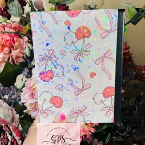 Peut inclure: Un cahier avec une couverture orn&eacute;e d'un motif de cerises roses, de n&oelig;uds et de c&oelig;urs sur un fond holographique. Le cahier a un dos noir et le texte "You dream it we make it gps creations".