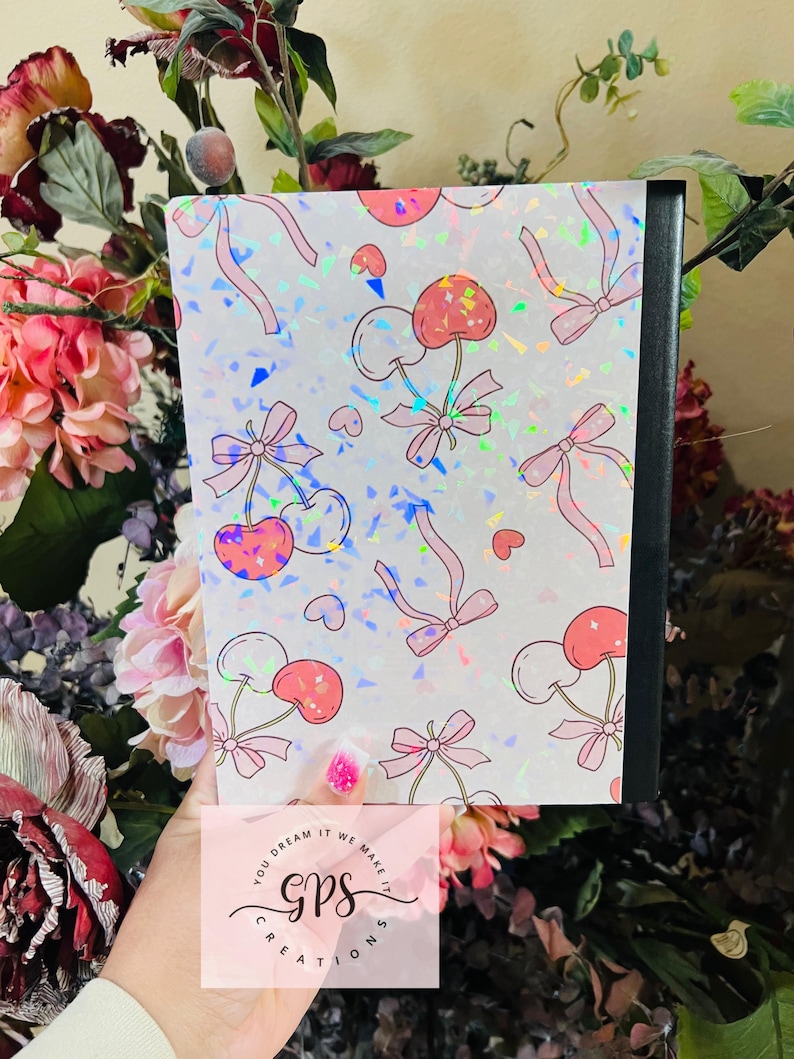 Peut inclure: Un cahier avec une couverture orn&eacute;e d'un motif de cerises roses, de n&oelig;uds et de c&oelig;urs sur un fond holographique. Le cahier a un dos noir et le texte "You dream it we make it gps creations".