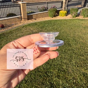 May include: A clear, plastic bottle stopper with a spiral design. The stopper is held in a hand against a background of green grass and a residential neighborhood. The text "YOU DREAM IT WE MAKE IT GPS CREATIONS" is visible.