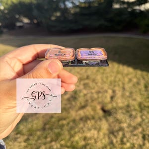 May include: Two small, rectangular, decorative items with a metallic border. One is light brown with the letter "L", the other is purple with the letter "R". The items are on a black base. The background is a green lawn and trees.