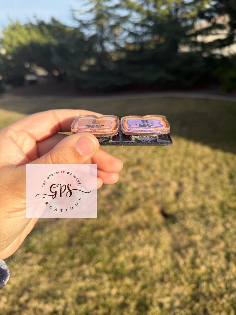 May include: Two small, rectangular, decorative items with a metallic border. One is light brown with the letter "L", the other is purple with the letter "R". The items are on a black base. The background is a green lawn and trees.