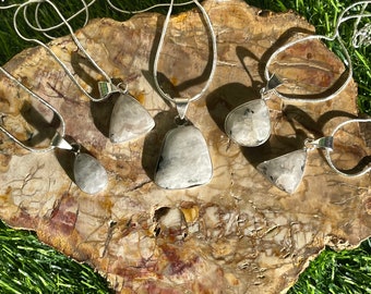 May include: Five silver necklaces with different shaped pendants made of white and gray stone. The necklaces are displayed on a piece of petrified wood.