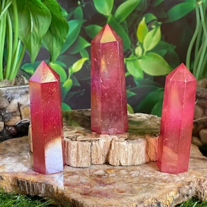 May include: Three pink aura quartz crystal points on a wooden slab, set against a backdrop of green foliage. The crystals have a gradient of colors, from a light pink at the base to a vibrant pink and gold at the top.