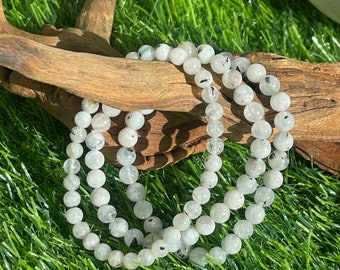 May include: Three white gemstone bracelets with black flecks, stacked on a piece of driftwood. The bracelets are made of round beads and are lying on a bed of green grass.