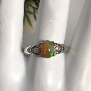 May include: A silver ring with an oval-shaped opal gemstone. The opal has a green, orange, and red iridescent sheen.