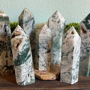 May include: Seven green and white agate crystal points with a natural, raw finish. The points are arranged in a row on a wooden surface.