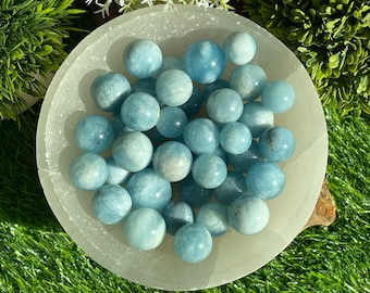 May include: A bowl filled with smooth, light blue aquamarine crystal spheres. The spheres are various sizes and are arranged in a random pattern. The bowl is white and has a smooth, polished surface.
