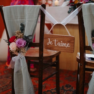 Puede incluir: Dos sillas de madera decoradas para una boda. Cada silla tiene una tela verde salvia drapeada sobre el respaldo, adornada con un arreglo floral de rosas rojas, rosas y moradas. Un letrero de madera con "Je t'aime" est&aacute; colgado en una silla.
