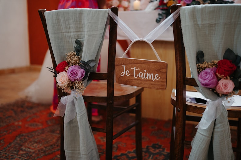 Puede incluir: Dos sillas de madera decoradas para una boda. Cada silla tiene una tela verde salvia drapeada sobre el respaldo, adornada con un arreglo floral de rosas rojas, rosas y moradas. Un letrero de madera con "Je t'aime" est&aacute; colgado en una silla.