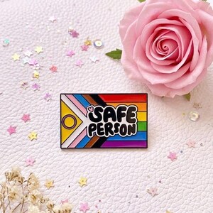 Peut inclure: Épinglette rectangulaire émaillée avec les mots "SAFE PERSON" en lettres noires, sur un fond arc-en-ciel. L'épinglette comprend des symboles représentant diverses identités LGBTQ+. Une rose rose et des confettis en forme d'étoiles entourent l'épinglette.