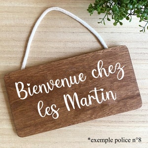 Puede incluir: Un letrero rectangular de madera con el texto "Bienvenue chez les Martin" en escritura blanca. El letrero tiene una cuerda blanca para colgar y un patr&oacute;n de vetas de madera. El texto "*exemple police n&deg;8" est&aacute; en la parte inferior.