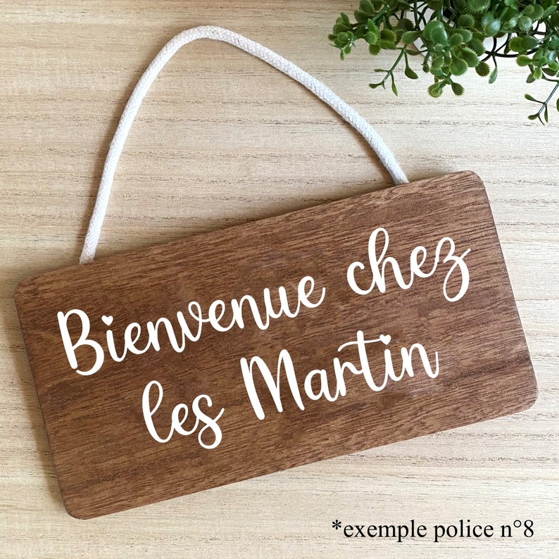 Puede incluir: Un letrero rectangular de madera con el texto "Bienvenue chez les Martin" en escritura blanca. El letrero tiene una cuerda blanca para colgar y un patr&oacute;n de vetas de madera. El texto "*exemple police n&deg;8" est&aacute; en la parte inferior.