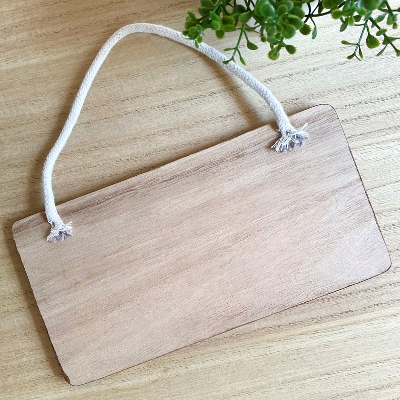 Puede incluir: Un letrero rectangular de madera con un acabado marr&oacute;n claro y una cuerda blanca para colgar. El letrero tiene esquinas redondeadas y un aspecto natural de veta de madera. La cuerda est&aacute; sujeta en la parte superior.