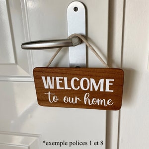 Puede incluir: Un letrero rectangular de madera con las palabras "WELCOME to our home" en letras blancas, colgado de un tirador de puerta. El letrero tiene un acabado de veta de madera marr&oacute;n claro y est&aacute; suspendido por una cuerda beige.