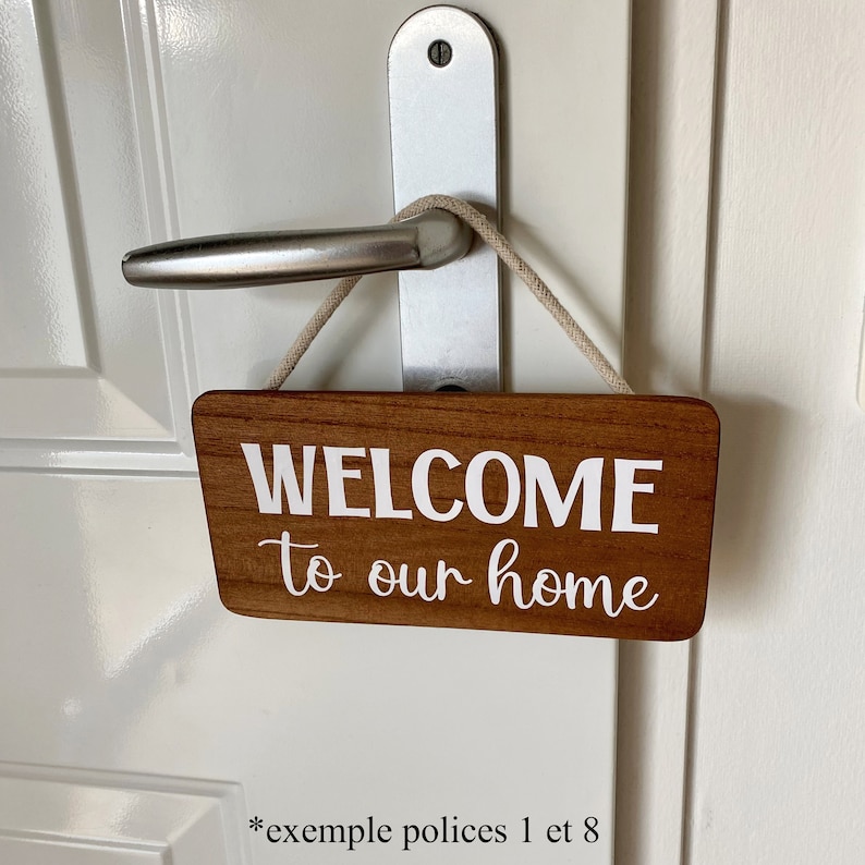 Puede incluir: Un letrero rectangular de madera con las palabras "WELCOME to our home" en letras blancas, colgado de un tirador de puerta. El letrero tiene un acabado de veta de madera marr&oacute;n claro y est&aacute; suspendido por una cuerda beige.
