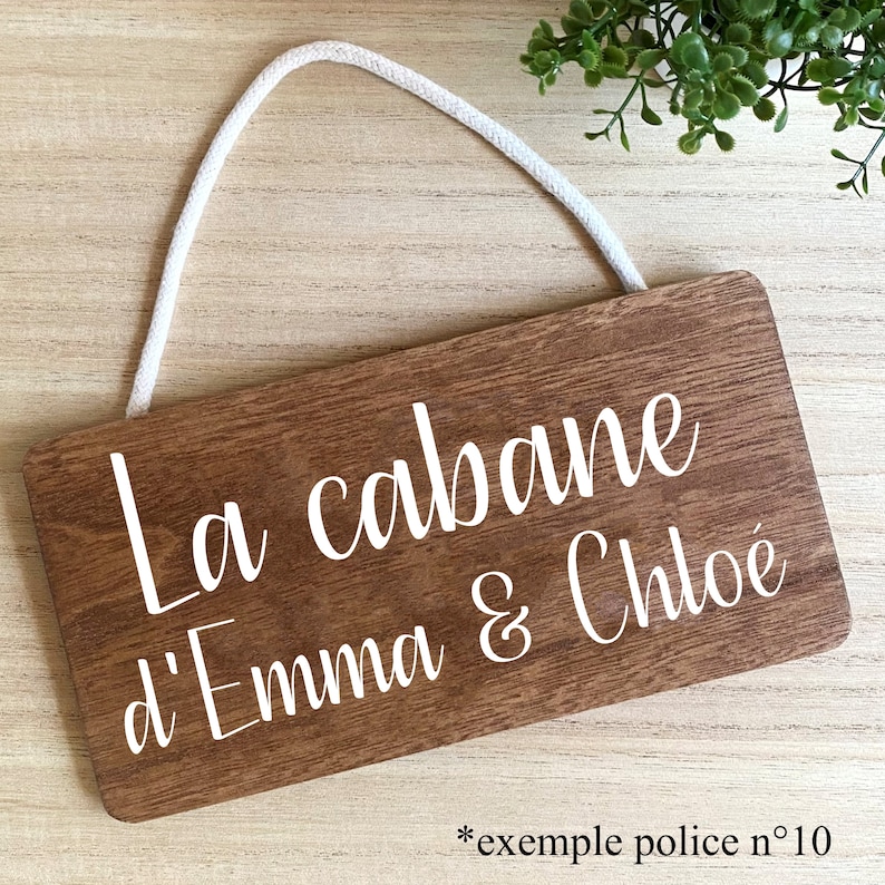 Puede incluir: Un letrero rectangular de madera con el texto "La cabane d'Emma & Chloe" en escritura blanca. El letrero tiene un acabado de veta de madera marr&oacute;n claro y una cuerda blanca para colgar.