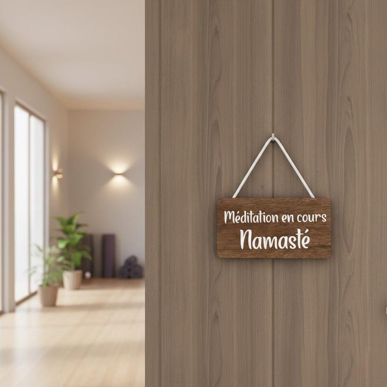 Puede incluir: Un letrero de madera con las palabras "M&eacute;ditation en cours" y "Namast&eacute;" en letras blancas cuelga de una puerta de madera. El letrero est&aacute; suspendido por un cord&oacute;n blanco. El fondo muestra un estudio de yoga.