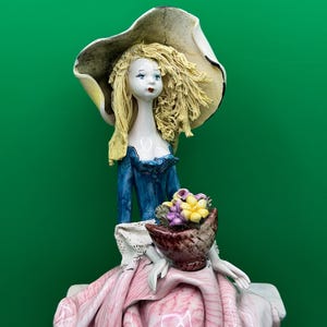 May include: A ceramic figurine of a woman wearing a large hat and a blue dress. The figurine holds a basket of flowers. The woman's dress is pink and white. The figurine has blonde hair and is set against a green background.
