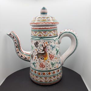 May include: A ceramic coffee pot featuring a hand-painted design of a deer surrounded by colourful floral and leaf motifs. The pot has a tall cylindrical body, a curved spout, a sturdy handle, and a matching lid with a pointed finial top.