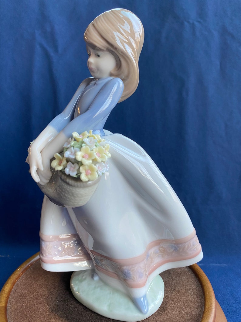 May include: A porcelain figurine of a girl in a blue dress and white skirt holding a basket of yellow and white flowers. The figurine is standing on a green base and is part of a musical figurine.