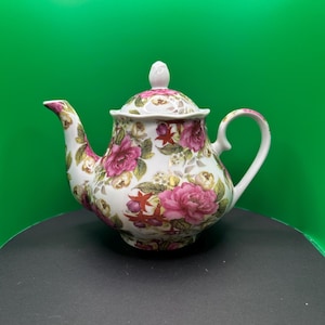 May include: A white porcelain teapot with a lid, adorned with a floral pattern featuring pink roses and green foliage. The teapot has a curved spout and handle, and a small knob on the lid.