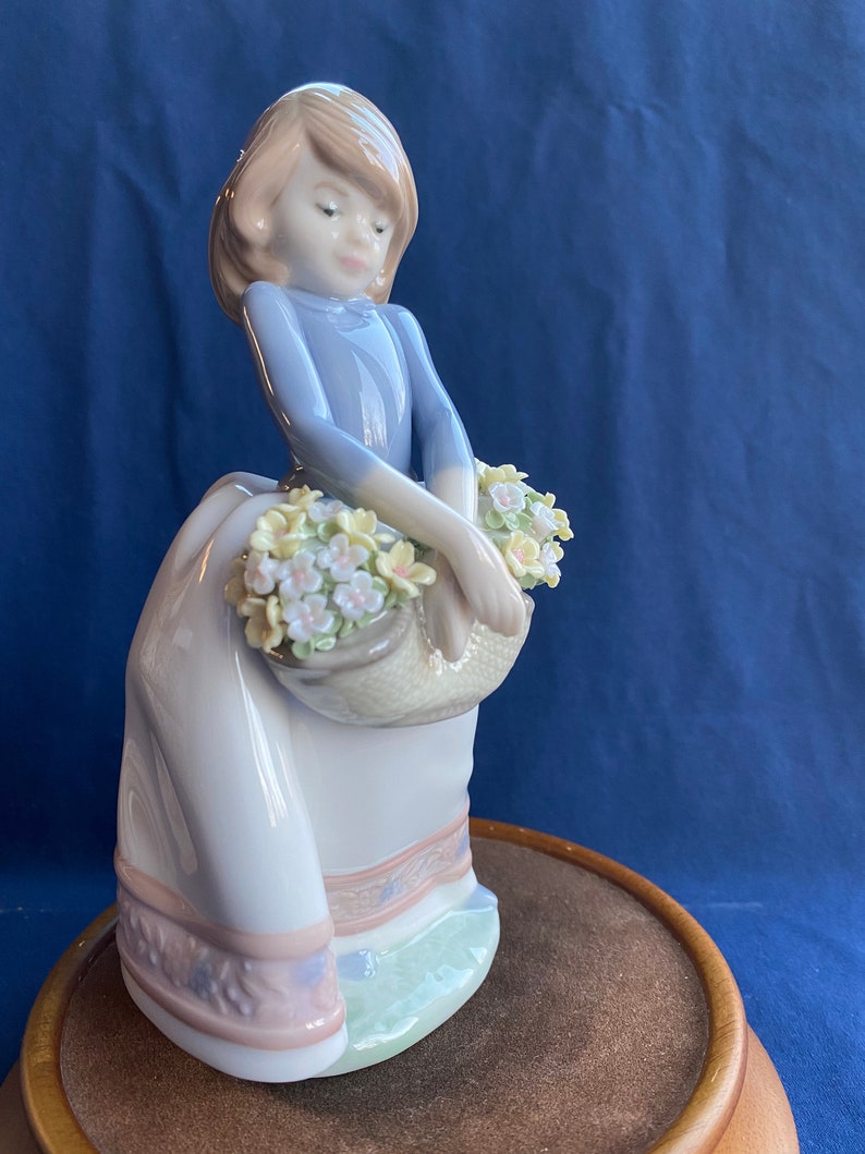 May include: A porcelain figurine of a girl in a blue dress and white skirt holding a basket of flowers. The figurine is standing on a wooden base.