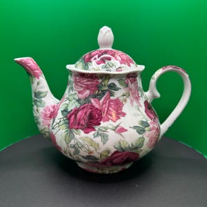 May include: A white porcelain teapot with a lid and handle, decorated with a floral rose pattern in shades of pink and red. The teapot has a curved spout and a white bud-shaped finial on the lid.