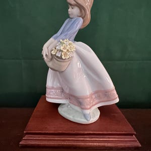 May include: A porcelain figurine of a girl in a blue and pink dress holding a basket of yellow flowers. The figurine is standing on a wooden base.