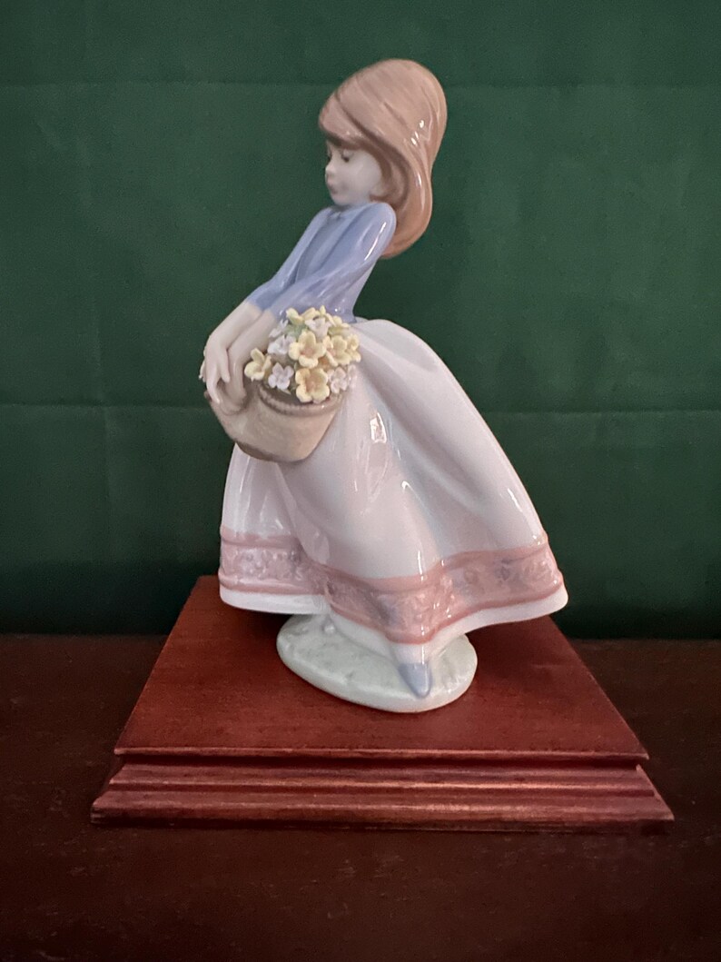 May include: A porcelain figurine of a girl in a blue and pink dress holding a basket of yellow flowers. The figurine is standing on a wooden base.
