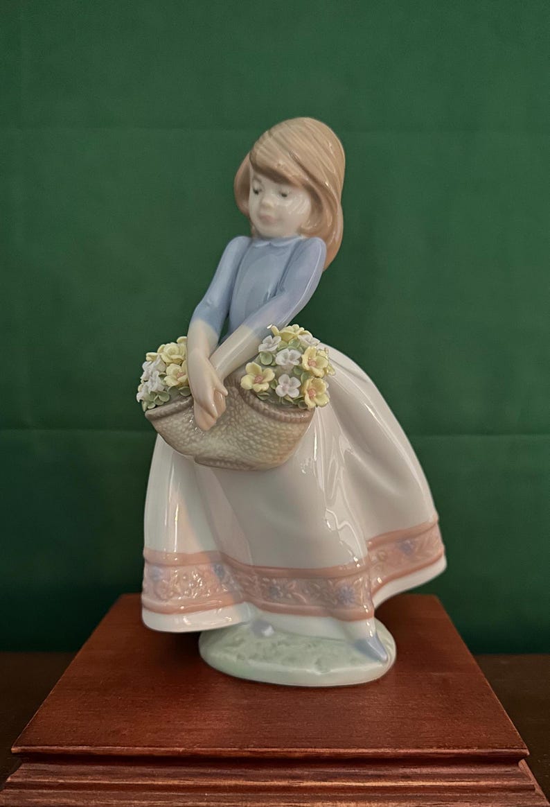 May include: A porcelain figurine of a girl in a blue dress and white skirt holding a basket of yellow and white flowers. The figurine is standing on a wooden base.