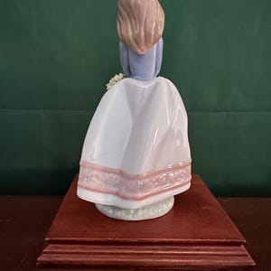 May include: A porcelain figurine of a girl with long brown hair wearing a white dress with pink trim and a blue top. She is holding a bouquet of flowers and is standing on a wooden base.