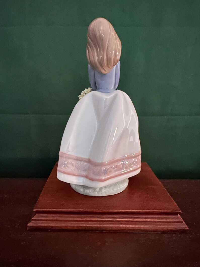May include: A porcelain figurine of a girl with long brown hair wearing a white dress with pink trim and a blue top. She is holding a bouquet of flowers and is standing on a wooden base.
