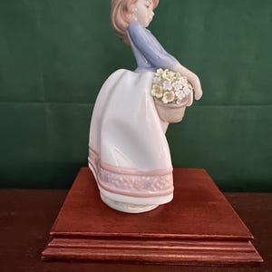 May include: A porcelain figurine of a girl in a blue dress and white skirt holding a basket of flowers. The figurine is on a wooden base.