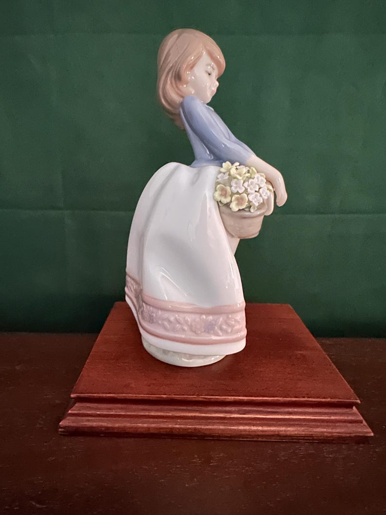 May include: A porcelain figurine of a girl in a blue dress and white skirt holding a basket of flowers. The figurine is on a wooden base.